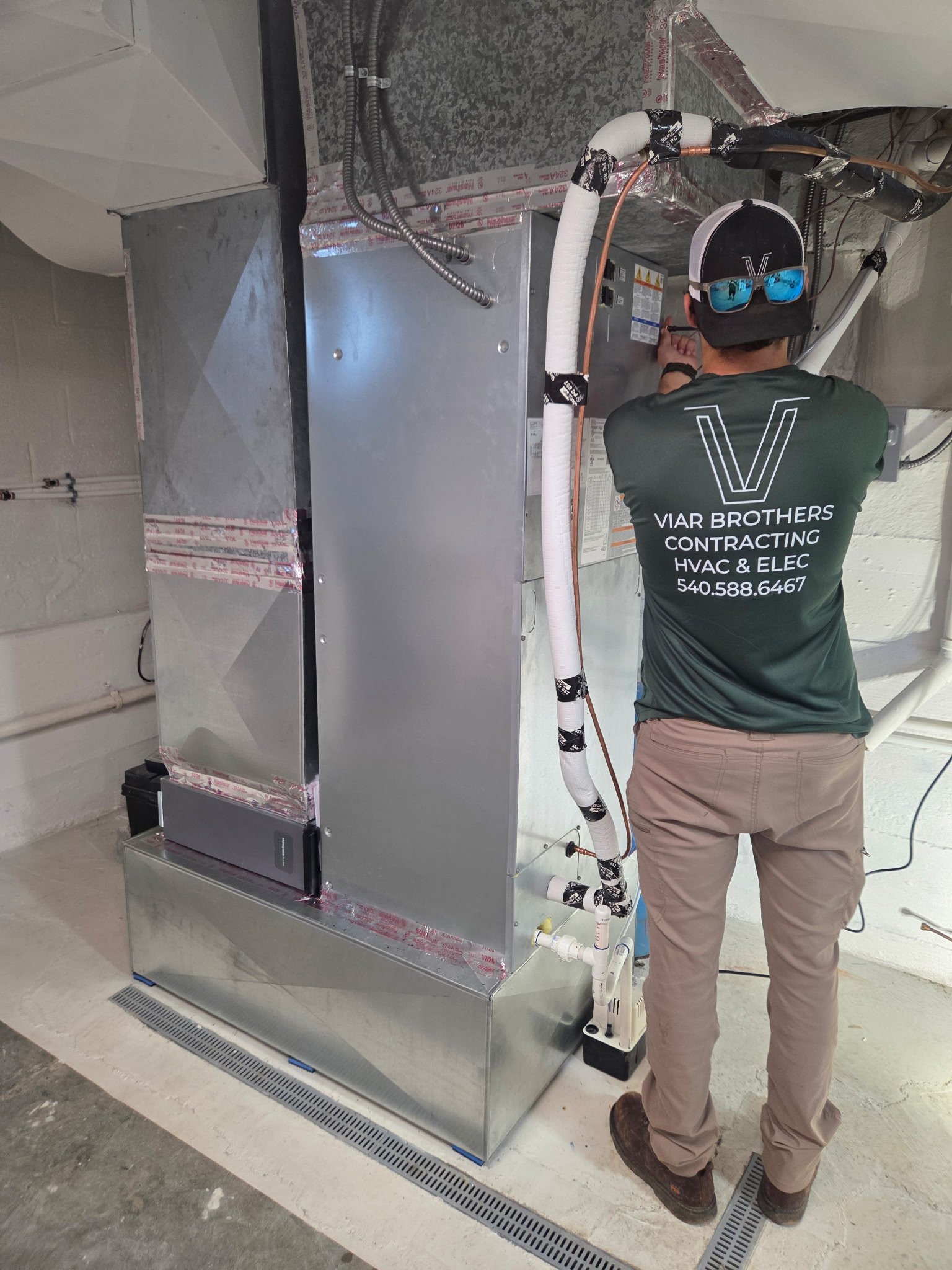 An HVAC technician in a green company uniform servicing a newly installed residential air handler and furnace unit inside a clean basement.